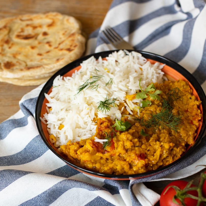 A bowl of Indian dal and white rice, garnished with herbs, next to flatbread and tomatoes.