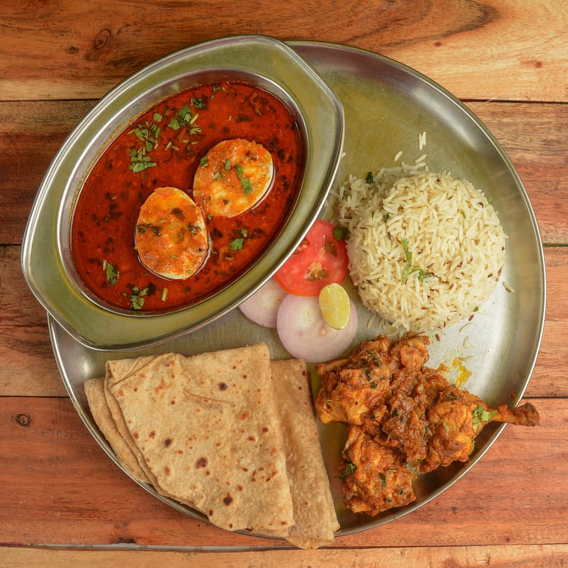 Indian thali with egg curry, chicken, rice, roti, and salad on a wooden table.